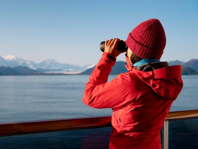 Alaska Glacier Bay cruise ship passenger looking at Alaskan mountains with binoculars exploring Glacier Bay National Park, just like Umbraco CMS users can explore software updates. 