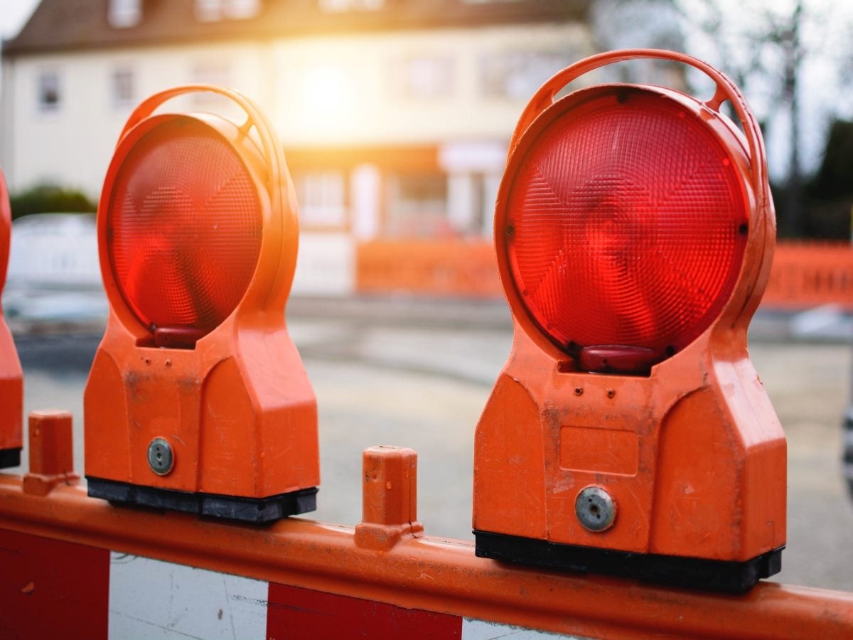 A close-up photograph of two bright orange road construction warning lights mounted on a striped traffic barricade, with a residential street and houses softly blurred in the background. The illuminated red beacons symbolize a roadblock, delay or obstacle, making the image suitable for stories about business challenges, stalled projects or organizational barriers.