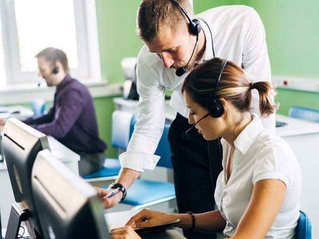 Contact center operators at work wearing headsets and using computers with one worker leaning in to help another in piece about the connection between contact centers and customer retention rates. 