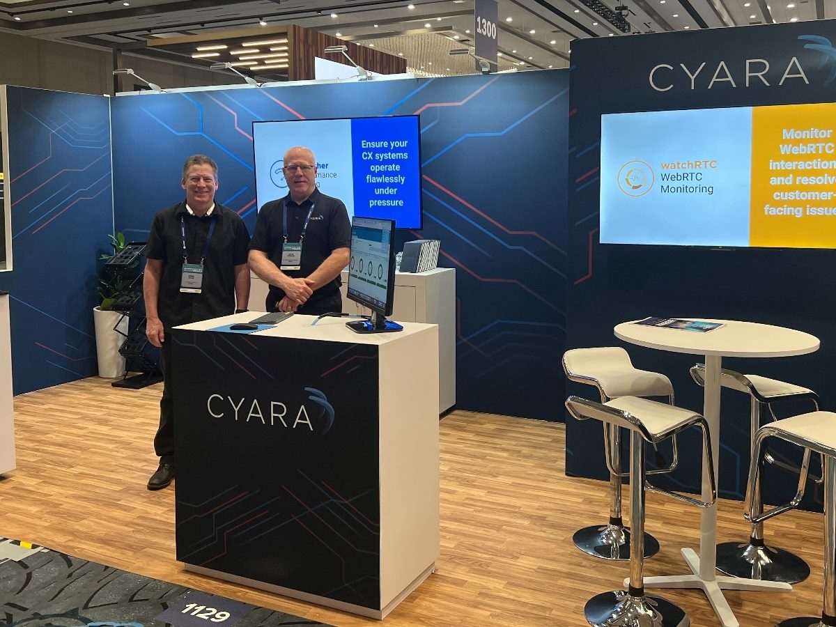 Two Cyara representatives stand at a trade show booth showcasing the company's CX validation and WebRTC monitoring solutions, with branded signage and a demo screen in the background.