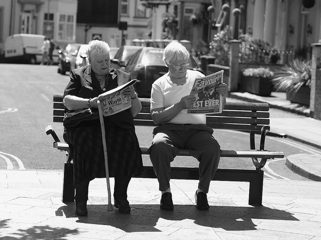 Woman and man sitting on a bench reading a newspaper in a black-and-white photo.