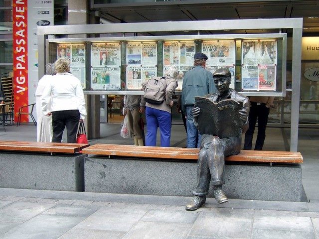 Statue of a man sitting down, legs crossed, reading a newspaper, in front of people looking into a glass case full of newspapers.