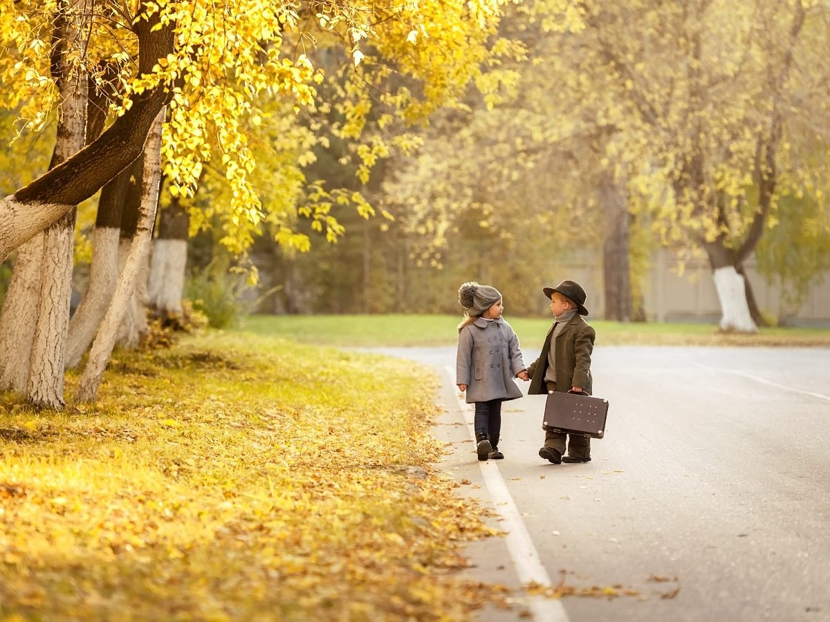 Two young children walk hand in hand along a quiet road lined with autumn trees. The trees have golden-yellow leaves, and the trunks are painted white at the base. One child wears a gray coat and knit hat, while the other wears a dark coat, scarf, and wide-brimmed hat, carrying a small vintage suitcase. The scene is warmly lit with a soft fall glow.