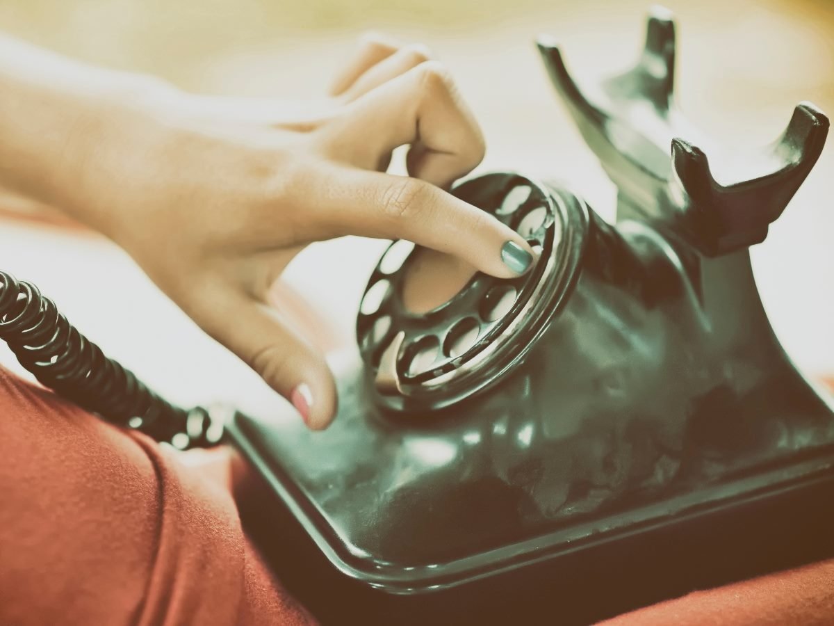 A close-up of a person dialing a vintage rotary telephone with a teal-painted fingernail.