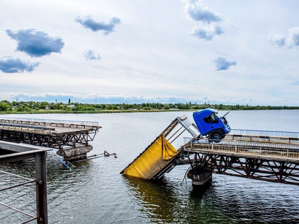 A blue semi-truck sits precariously on the broken edge of a collapsed bridge spanning a body of water. A large section of the bridge has fallen into the water below, leaving a wide gap between the remaining sections. The scene is set under a cloudy sky, with shoreline vegetation and distant buildings visible in the background.