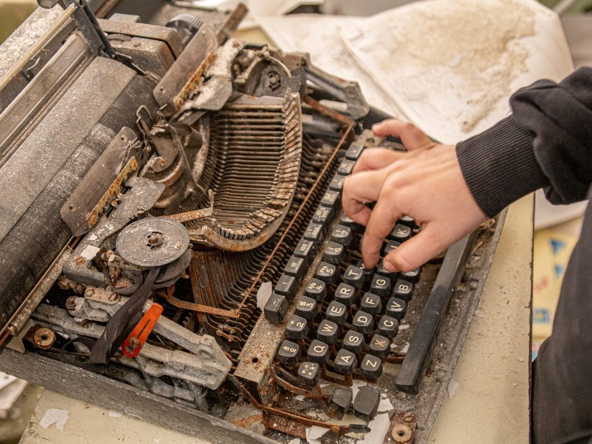 A close-up, high-angle photo shows a person's hands typing on a heavily rusted, dust-covered vintage typewriter. The exposed type-bar mechanism sits open and disassembled at the center of the frame, its metal arms coated in orange rust and gray dust. Only one hand is visible pressing down on the worn black keys, wearing a dark ribbed sweater cuff. Yellowed, stained paper sits in the background near the machine's carriage, and loose paper scraps and grit cover the surface below.