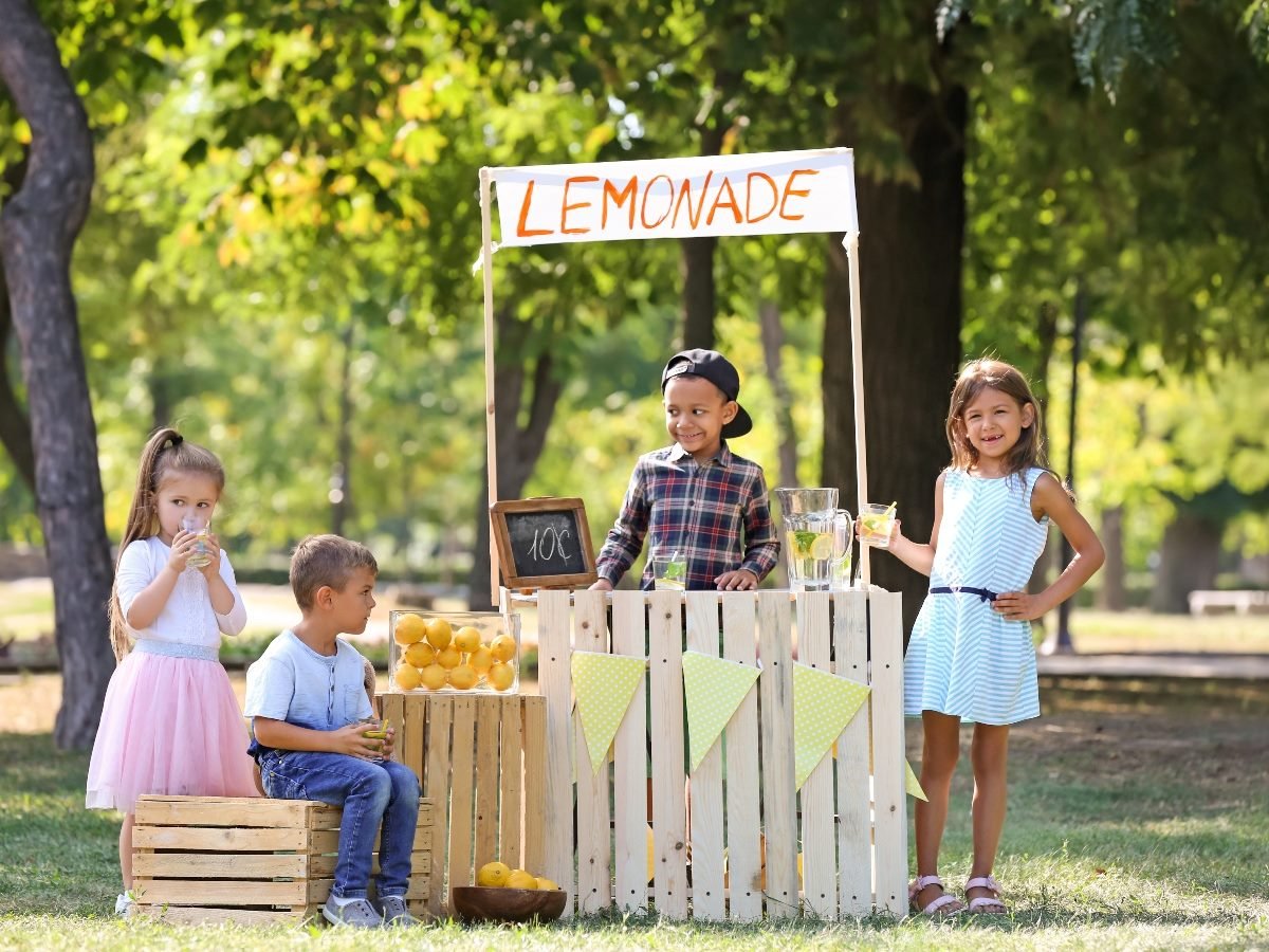 A group of young children run a lemonade stand outdoors on a sunny day. Two girls stand on either side holding cups of lemonade, while two boys sit and stand near crates filled with lemons. A hand-painted “Lemonade” sign hangs above the wooden stand, and pitchers of lemonade sit on the counter. Trees and greenery fill the background.