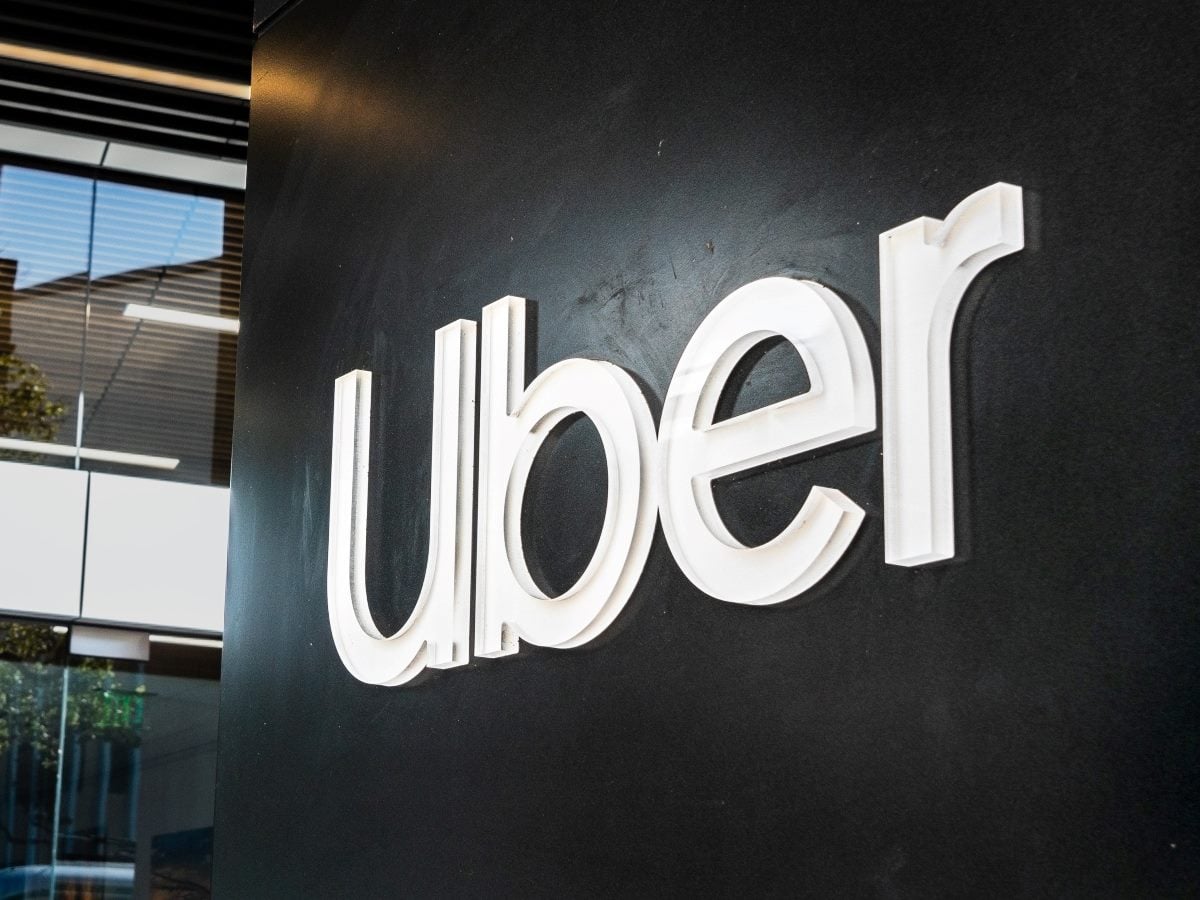 Close-up of an illuminated Uber logo mounted on a black interior wall inside an office building. Glass windows, modern lighting and architectural details are visible in the background, highlighting the company's corporate workspace.