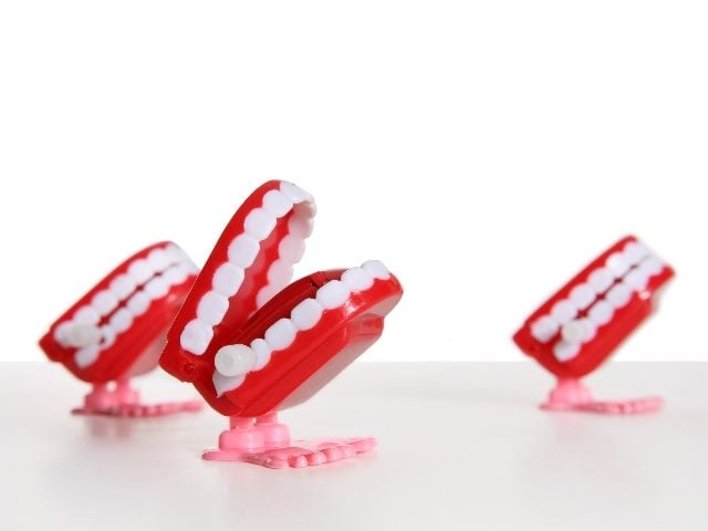 Three chattering teeth toys with red gums and pink feet sit on a white surface with a white background, suggesting the history of chatbots.