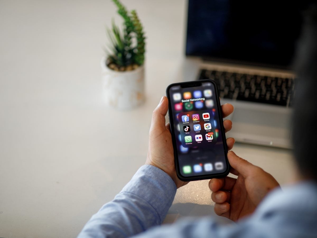 A person seated at a desk holds a smartphone displaying an open "Social Networking" folder with app icons for Facebook, Instagram, YouTube, TikTok, Twitter, Google, WhatsApp, Flickr, and Gmail. The person wears a light blue button-down shirt, with a laptop and a small succulent plant in a white marble-patterned pot visible blurred in the background on a light desk surface.