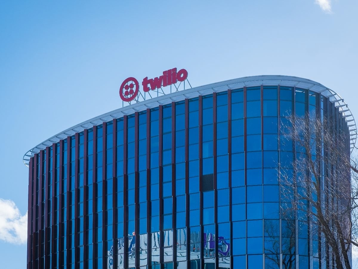 Modern glass office building in Tallinn, Estonia, with the Twilio logo mounted on the rooftop. The curved blue reflective facade is framed by leafless trees under a bright clear sky. Twilio is an American cloud communications platform company.