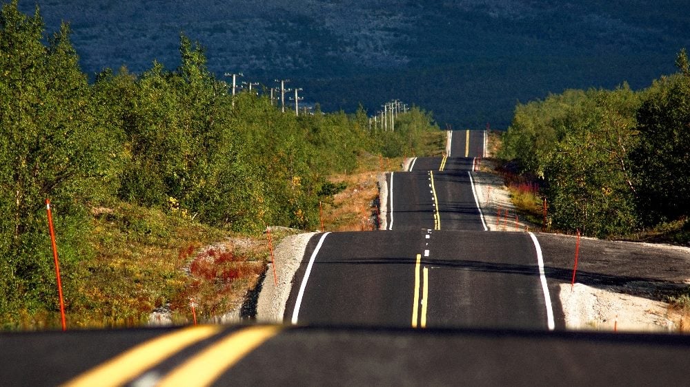 A bumpy, hilly road is seen disappearing into the horizon with trees and greenery visible alongside the road as well as some electrical poles in piece about customer onboarding regulatory compliance.