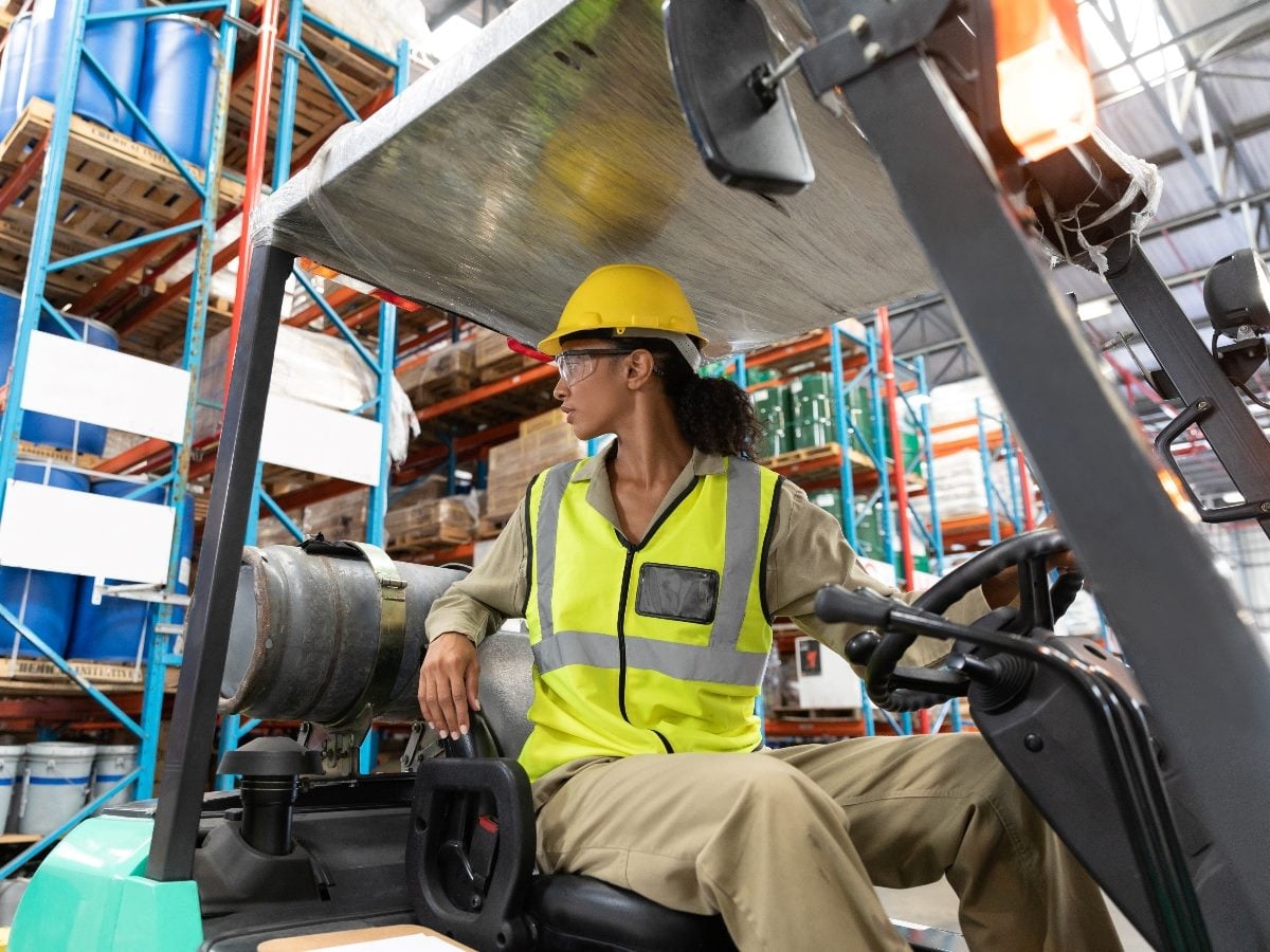 Warehouse worker wearing a yellow hard hat and high-visibility vest operates a forklift inside a distribution center, with industrial shelving and stacked barrels visible in the background.