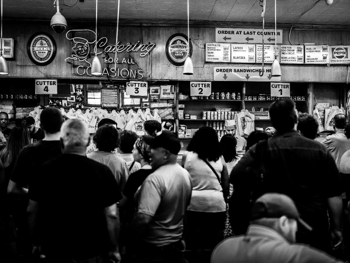 Customers line up at a busy deli counter beneath retro signage reading “Catering for All Occasions,” as servers take sandwich orders behind the counter in a black-and-white photo.