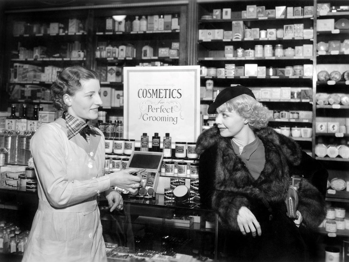 A black-and-white cosmetics shop scene, probably from the 1930s, showing a female store clerk demonstrating a makeup compact to a stylish woman wearing a fur coat and hat. Shelves filled with beauty products line the background, and a sign reading “Cosmetics for Perfect Grooming” sits between them.
