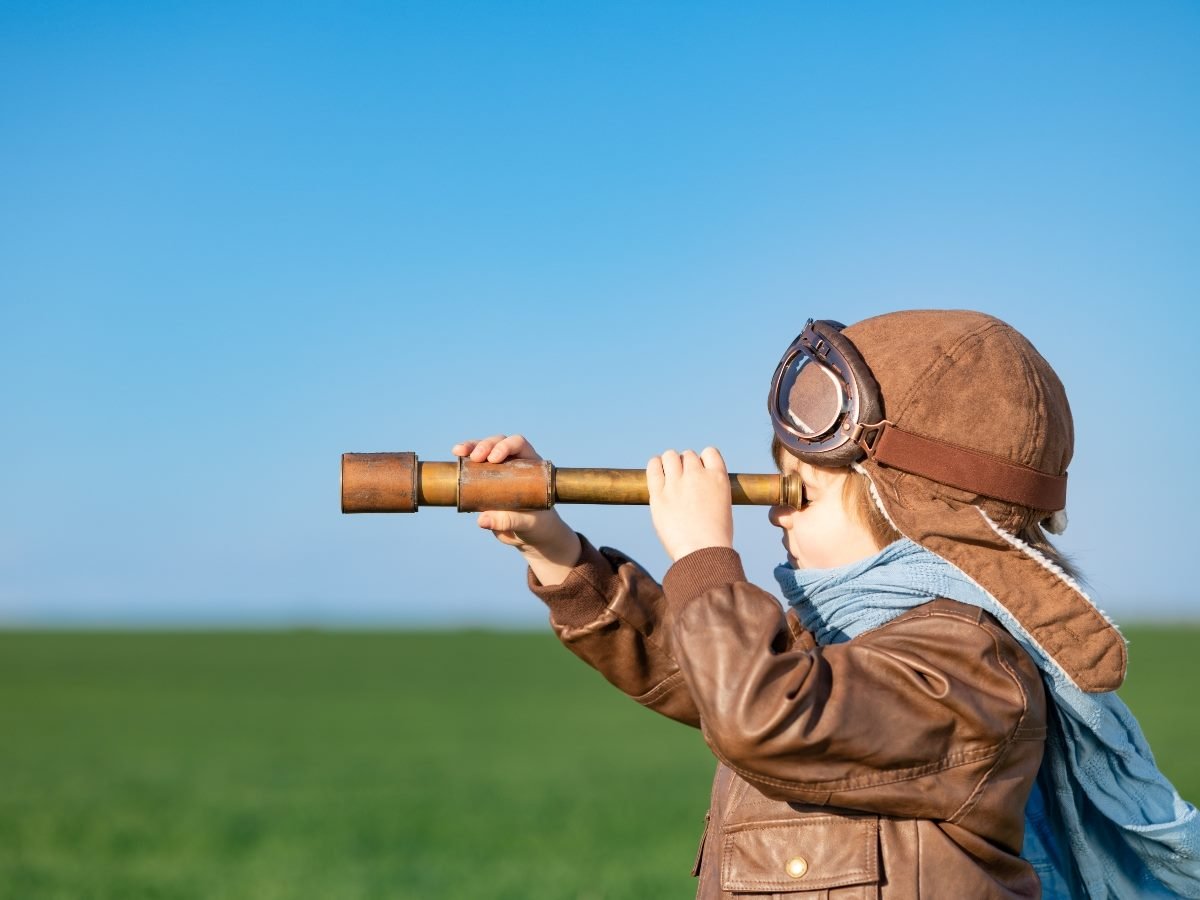 A young child dressed as an aviator, wearing a leather jacket, goggles, and a scarf, looks through a brass telescope across a green field under a clear blue sky.