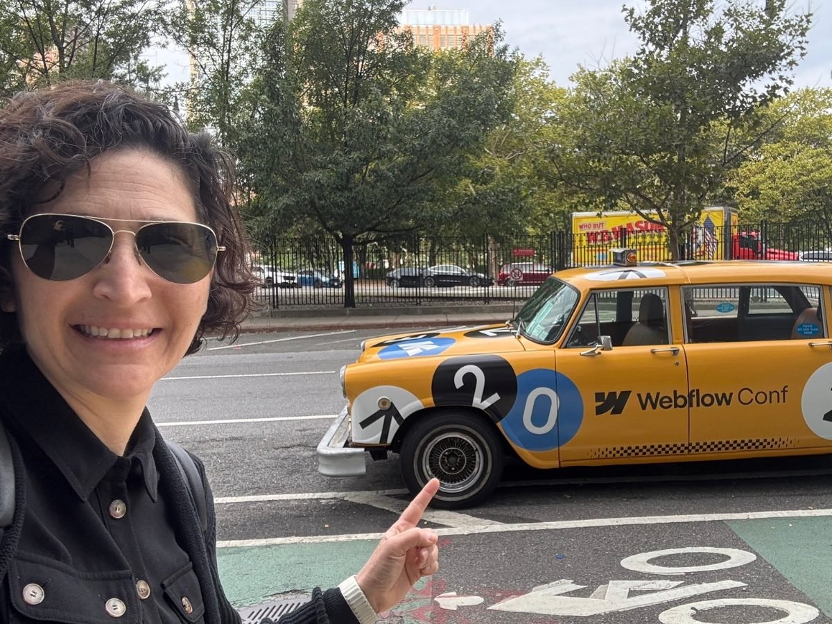 A person smiles and points toward a yellow car decorated with “Webflow Conf” branding, parked along a city street.
