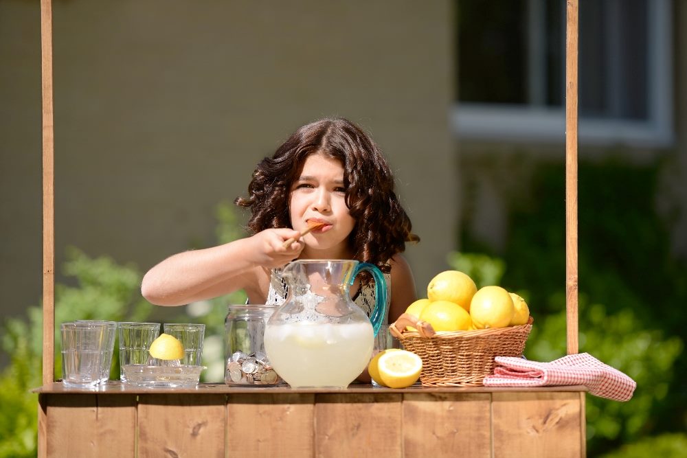 A child makes and tastes lemonade in a pitcher on a sunny day at a lemonade stand with a basket of lemons on her right and glasses on her left in piece about customer retention and customer churn.