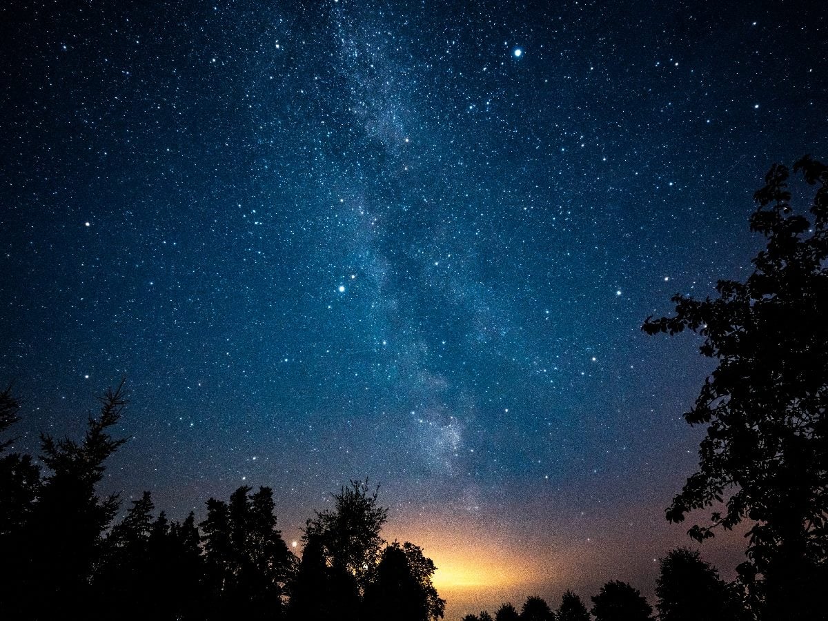 A star-filled night sky stretches above a dark tree-lined horizon, with the Milky Way visible as a bright vertical band across the center. A soft golden glow rises near the horizon, contrasting with the deep blue sky and silhouetted trees.