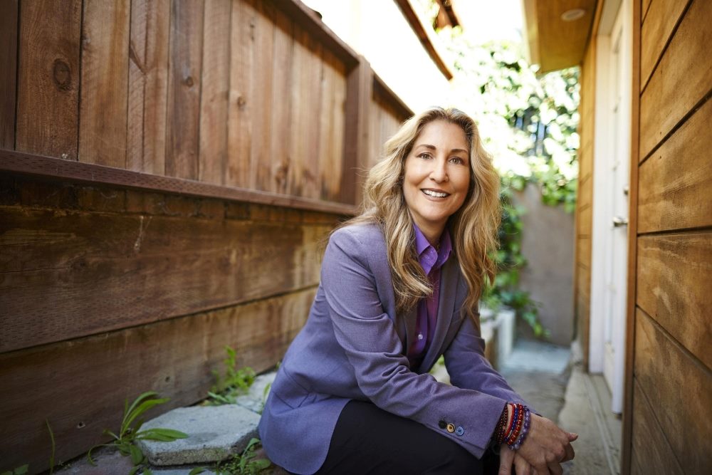 Tiffani Bova wears a purple suit jacket while sitting outside in a wooden walkway next to a house with rocks and greenery in the background in piece about the importance of CX and EX.