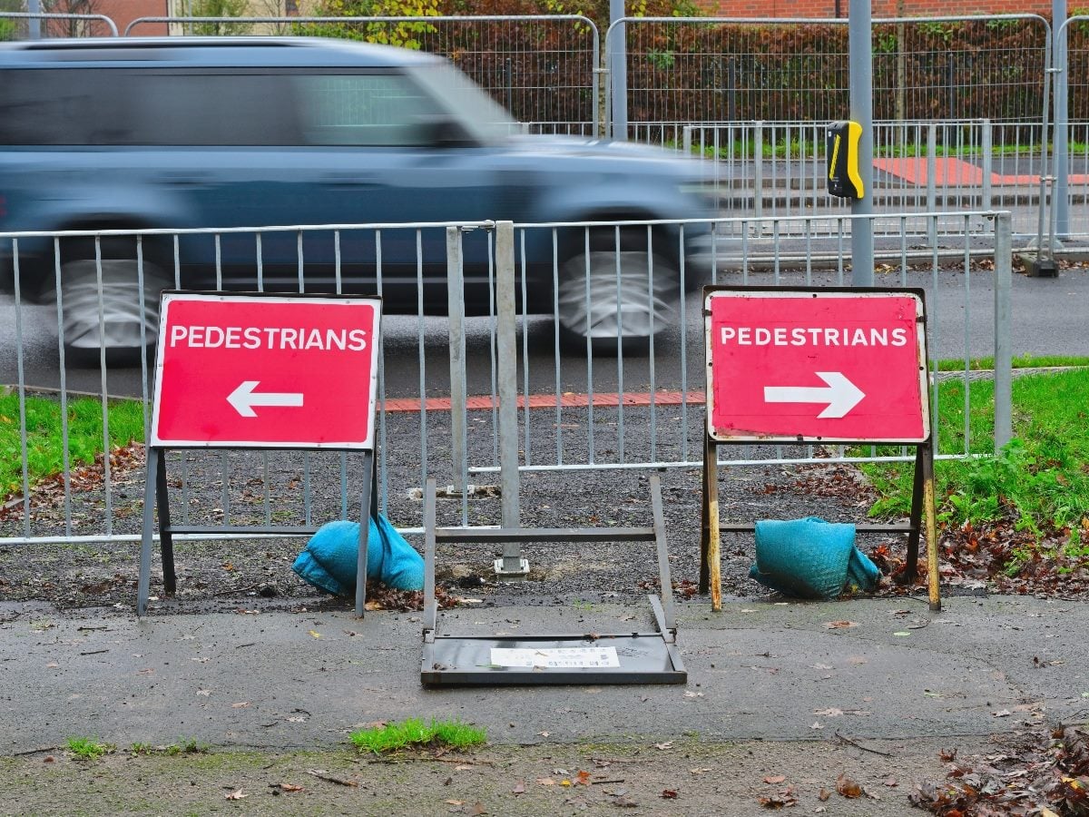 Two temporary pedestrian detour signs point in opposite directions beside a fenced roadside construction area as a blurred SUV passes in the background.