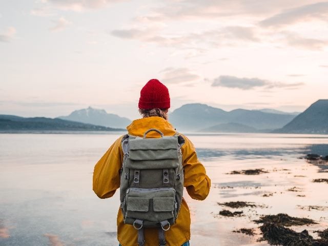 Back view of male tourist with rucksack and wearing a yellow coat and red knit cap standing on coast in front of mountains in the distance while journey, suggesting impact of journey orchestration.