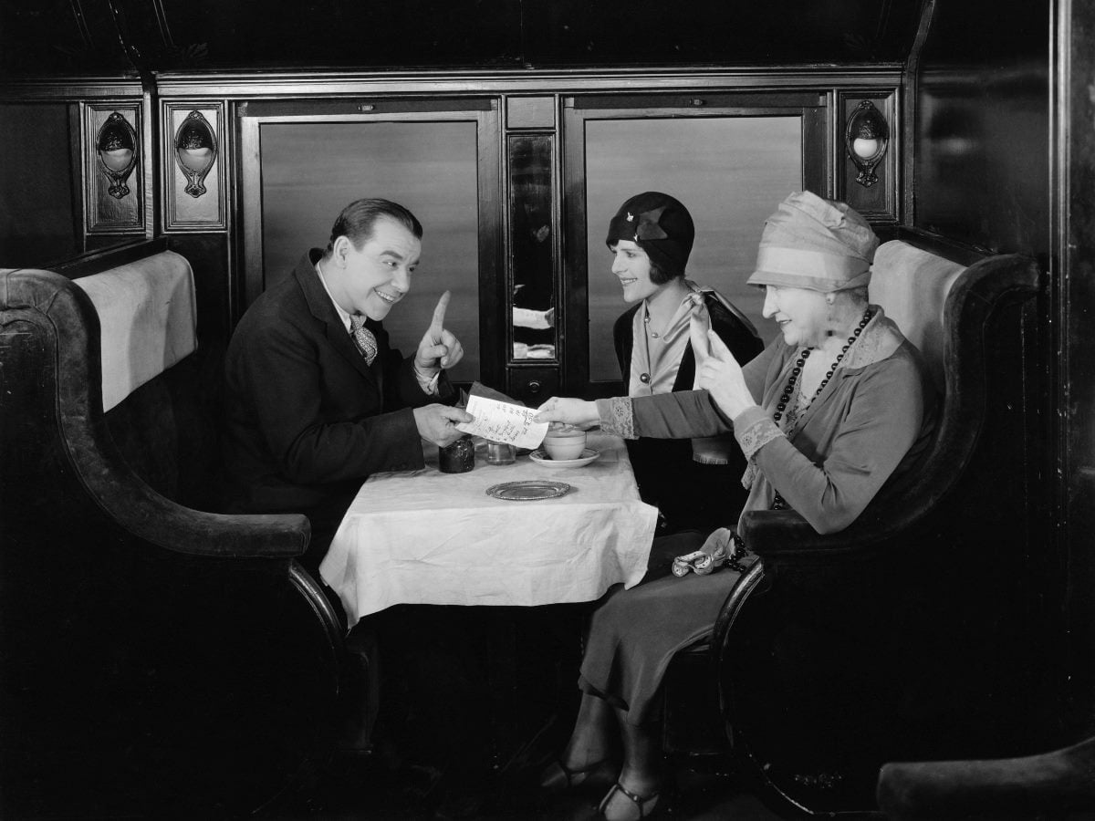 A man and two women share a conversation at a dining table aboard a train car, circa 1920s, in a scene suggesting attentive, personalized customer service.