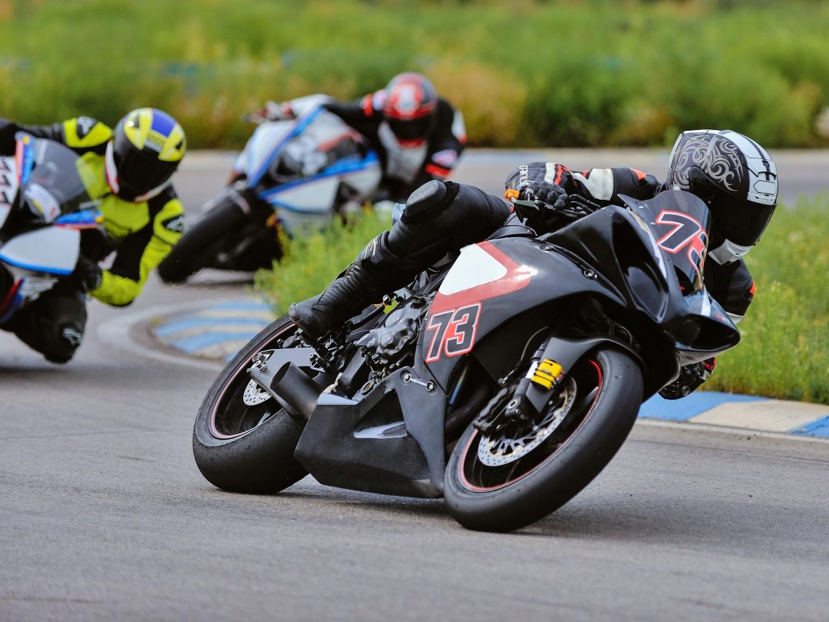 Motorcycle racers leaning into a sharp curve on a racetrack, with rider #73 in the lead wearing a black suit and helmet with silver patterns, followed closely by competitors in brightly colored gear.