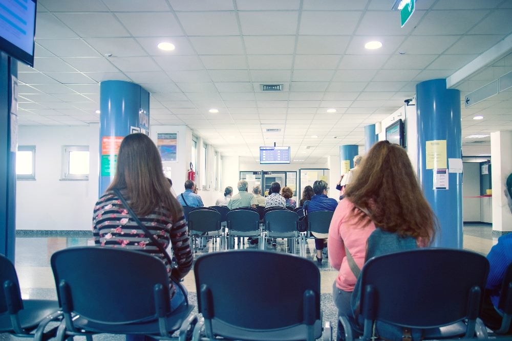People wait their turn in blue chairs in a hospital waiting room with blue support beams, suggesting the importance of understanding customer behavior.