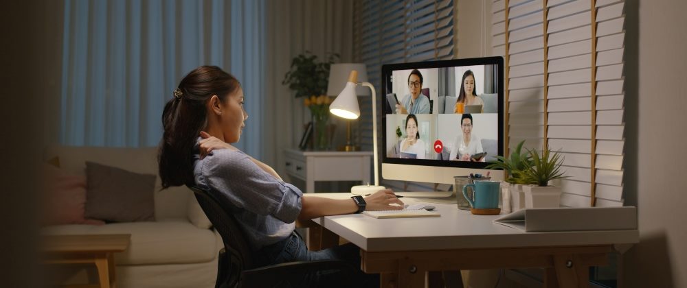 A woman sits in a darkened room attending an online meeting, symbolizing the need to avoid hybrid customer advisory board meetings.