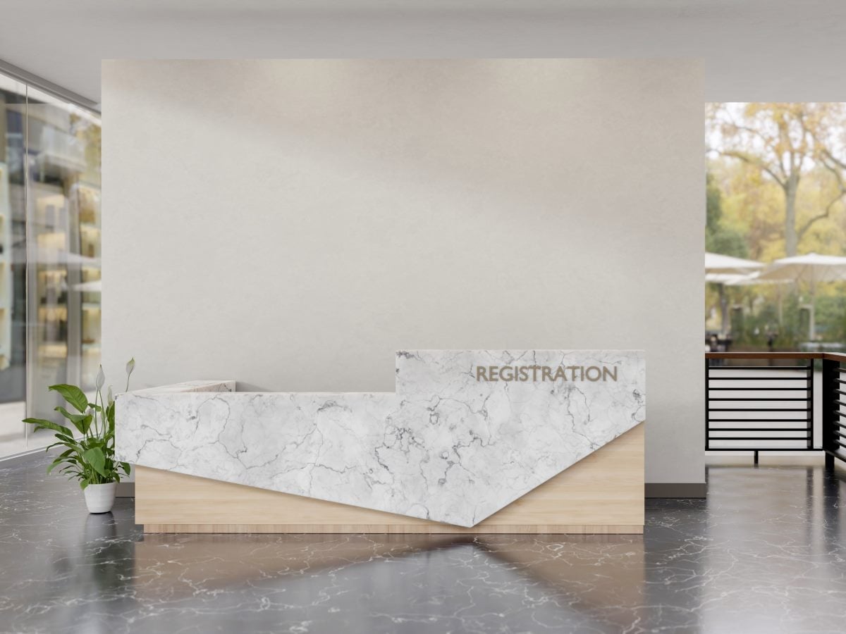 A modern hotel or office lobby reception desk with an angular, wedge-shaped front clad in white-and-gray veined marble over a light natural wood base, with "REGISTRATION" spelled out in raised bronze-toned letters on the taller marble panel. A leafy potted peace lily sits to the left of the desk on a polished dark terrazzo floor that reflects the surrounding space, in front of a large blank beige feature wall. Floor-to-ceiling glass on the left reveals a softly blurred exterior building facade, while an open archway on the right leads to an outdoor patio with cream umbrellas and autumn-colored trees, and a dark wood-and-metal railing partition is visible just inside that opening.