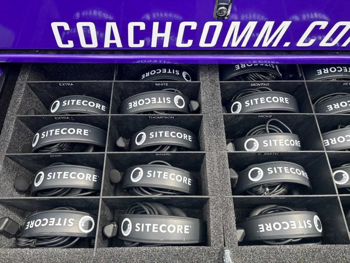 A set of black Sitecore-branded headsets neatly organized in a foam-lined case labeled with individual names and stored beneath a purple CoachComm compartment.