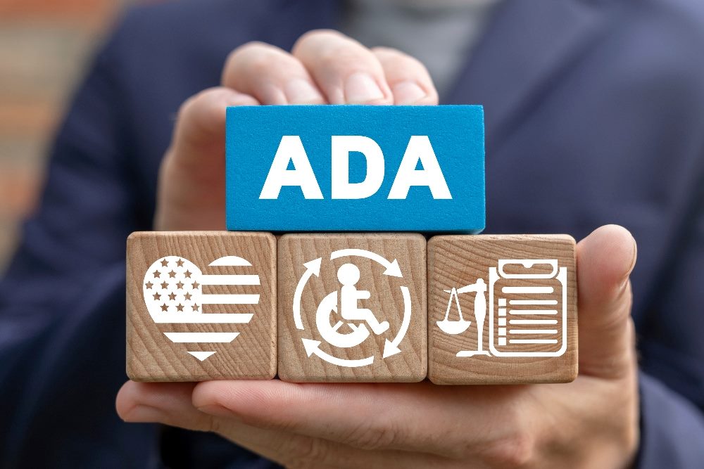 A person holds a long blue wooden block with the letters ADA for the Americans With Disabilities Act with three brown blocks underneath it depicting a heartshaped American flag, a wheelchair access symbol and scales of justice, in piece concerning accessible web design.