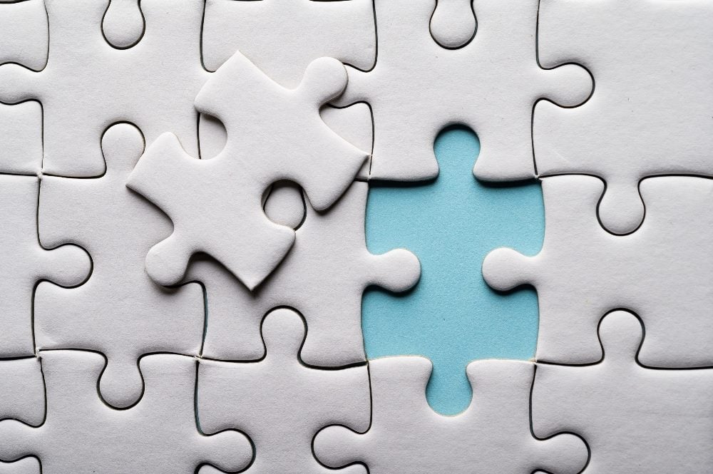 An image of a white jigsaw puzzle with one piece missing. The missing piece reveals a light blue background underneath, highlighting the empty space. A single puzzle piece is placed beside the gap, ready to fit into the empty spot. The overall image emphasizes the importance of each piece in completing the whole puzzle in piece about Mach architecture and composability.