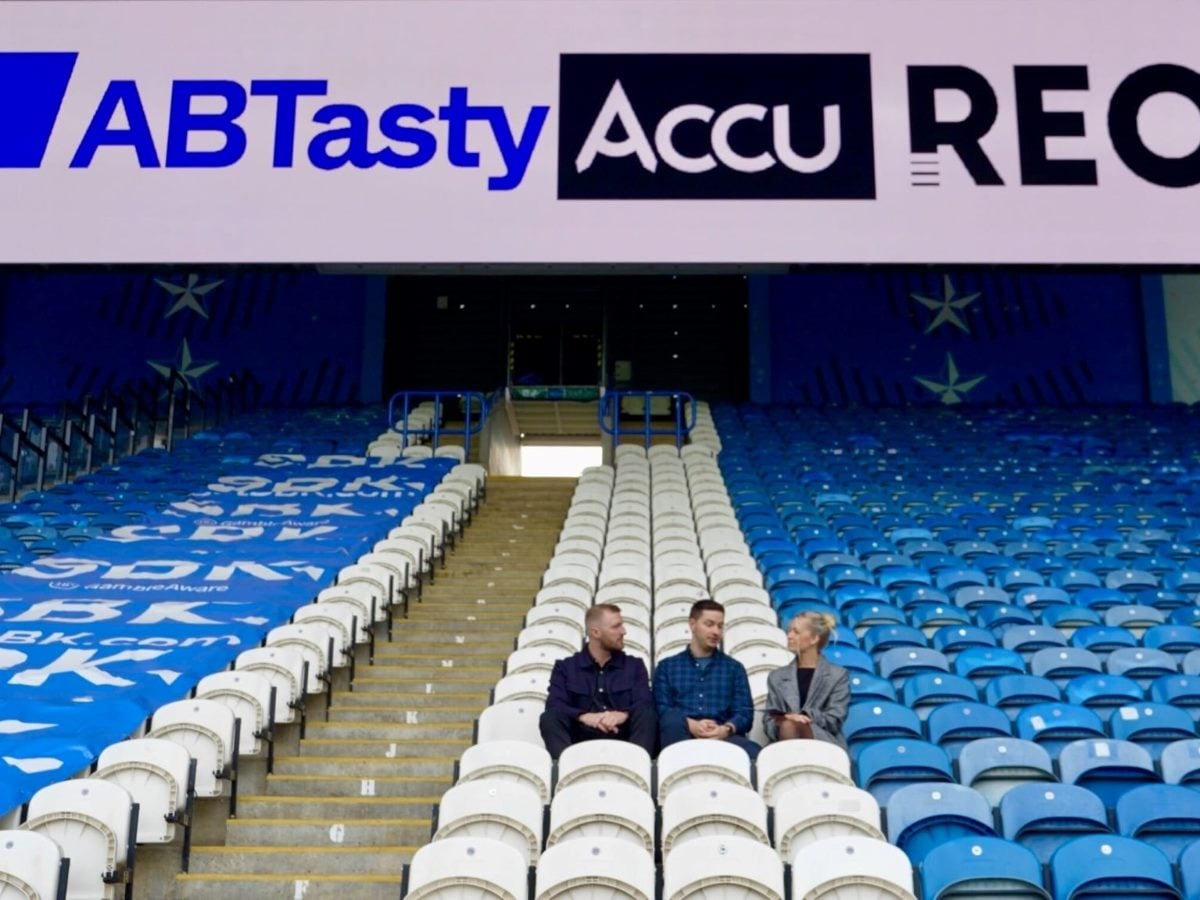 Three people sit and talk in mostly empty blue-and-white stadium seating beneath large overhead signage reading “AB Tasty” and “Accu…,” with rows of seats and a central stairway leading up behind them.