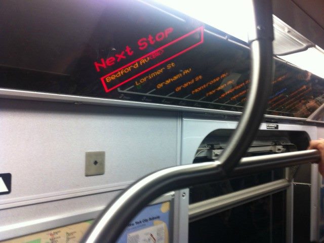 Neon red digital sign on ceiling of a train that says, "Next Stop" along with several other stops.
