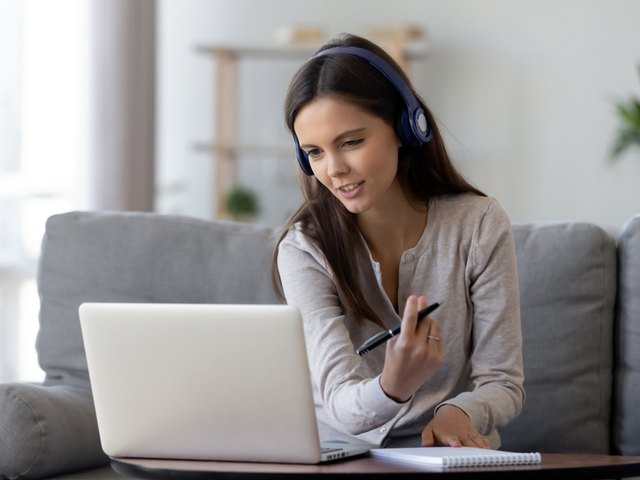 young businesswoman wearing headphones speaking and looking at her laptop while taking notes, in her living room.