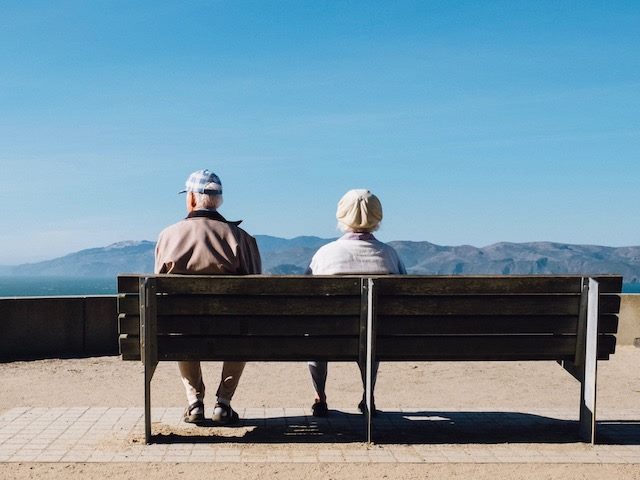 older couple sitting on a bench, with backs to the camera, looking out at nature 