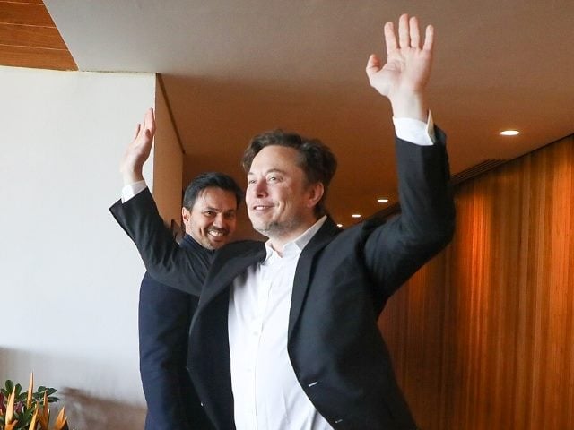 Elon Musk with his hands raised and smiling with an unidentified man behind him.